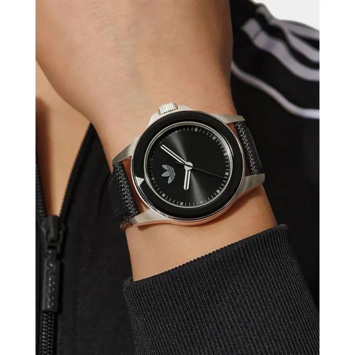 ADIDAS AOFH23016-5 Tuvplāns elegantam melnam rokas pulkstenim ar apaļu ciparnīcu, uz personas rokas, kas valkā melnu rāvējslēdzēja jaku.