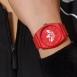 ADIDAS AOST23051-4 Lähivõte punasest käekellast silmapaistva logoga, kantud käel koos musta tõmblukukinnitusega jakiga.