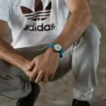 A close-up of a man in an Adidas shirt, squatting with his stylish blue watch prominently visible on his wrist.