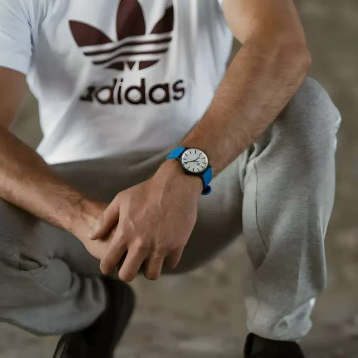 A close-up of a man in an Adidas shirt, squatting with his stylish blue watch prominently visible on his wrist.