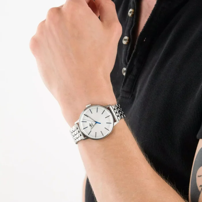 Stambiu planu matomas stilingas sidabrinis laikrodis su baltu ciferblatu ir mėlynomis rodyklėmis vienspalviame fone.