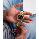 Stambiu planu užfiksuota ranka su auksiniu „Guess“ laikrodžiu juodu ciferblatu, laikoma atsipalaidavusi ant džinsinio audinio.