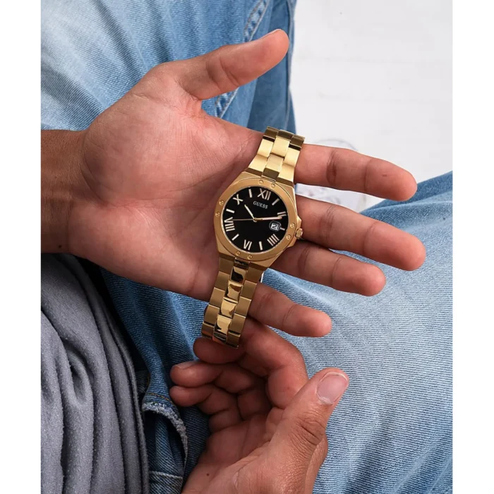 Stambiu planu užfiksuota ranka su auksiniu „Guess“ laikrodžiu juodu ciferblatu, laikoma atsipalaidavusi ant džinsinio audinio.