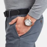 MONDAINE MH1.B1210.LG-2 Мужчина в сером свитере и синих брюках непринужденно держит руку в кармане, демонстрируя часы с коричневым кожаным ремешком на запястье.