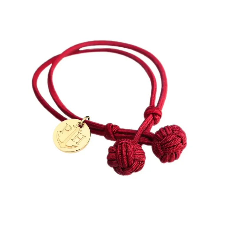 A red bracelet featuring three decorative knots and a gold pendant with initials and an anchor design.
