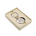 Elegantiškas sidabrinis raktų pakabukas su karūnos logotipu, elegantiškai įpakuotas į kreminės spalvos dovanų dėžutę.