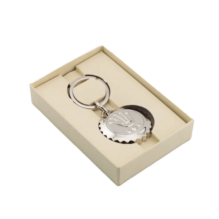 Elegantiškas sidabrinis raktų pakabukas su karūnos logotipu, elegantiškai įpakuotas į kreminės spalvos dovanų dėžutę.