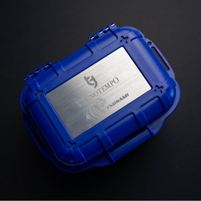Sininen suojakotelo, jossa hopeanvärinen metallikansi, jossa Tecnofempo- ja Tsunami-logot, mustaa taustaa vasten.