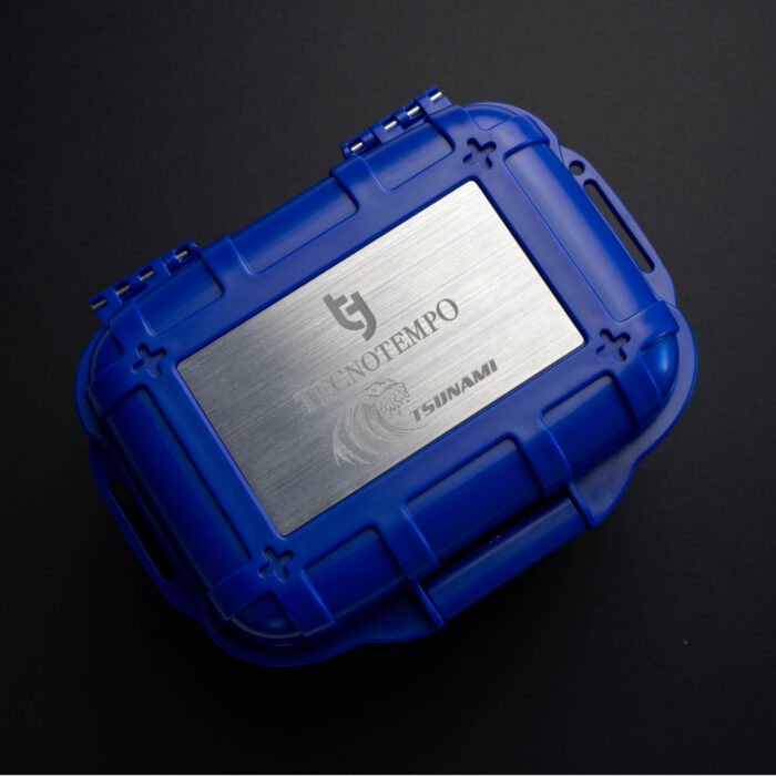 Mėlynas apsauginis dėklas su sidabro spalvos metaliniu dangteliu, ant kurio juodame fone pavaizduoti „Tecnofempo“ ir „Tsunami“ logotipai.