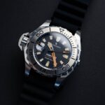 Lähikuva kestävästä sukeltajakellosta, jossa metallikuori, musta kumihihna ja tumma kellotaulu, jossa hehkuvat merkit ja oranssit yksityiskohdat.