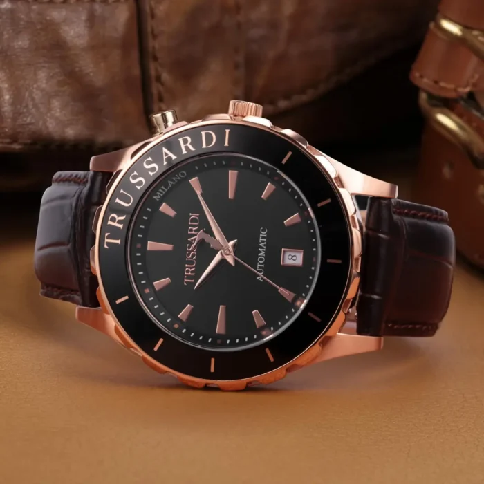 TRUSSARDI R2421143001-5 A stylish Trussardi automatic watch with a black dial, rose gold accents, and a brown leather strap, set against a textured background.