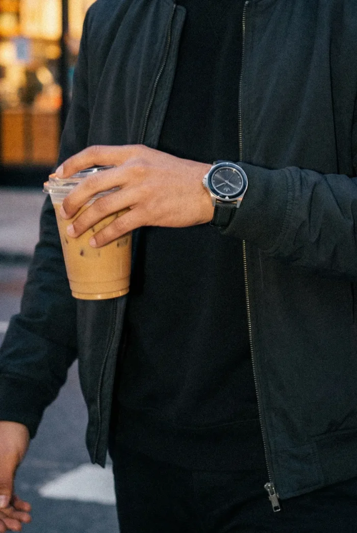 A person in a black jacket holds an iced coffee in a plastic cup. They wear a sleek watch and are walking on a city street with a blurred background.