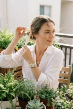 A woman in a white blouse ties her hair with a ribbon on a balcony. She's surrounded by green plants, exuding a calm and serene atmosphere.