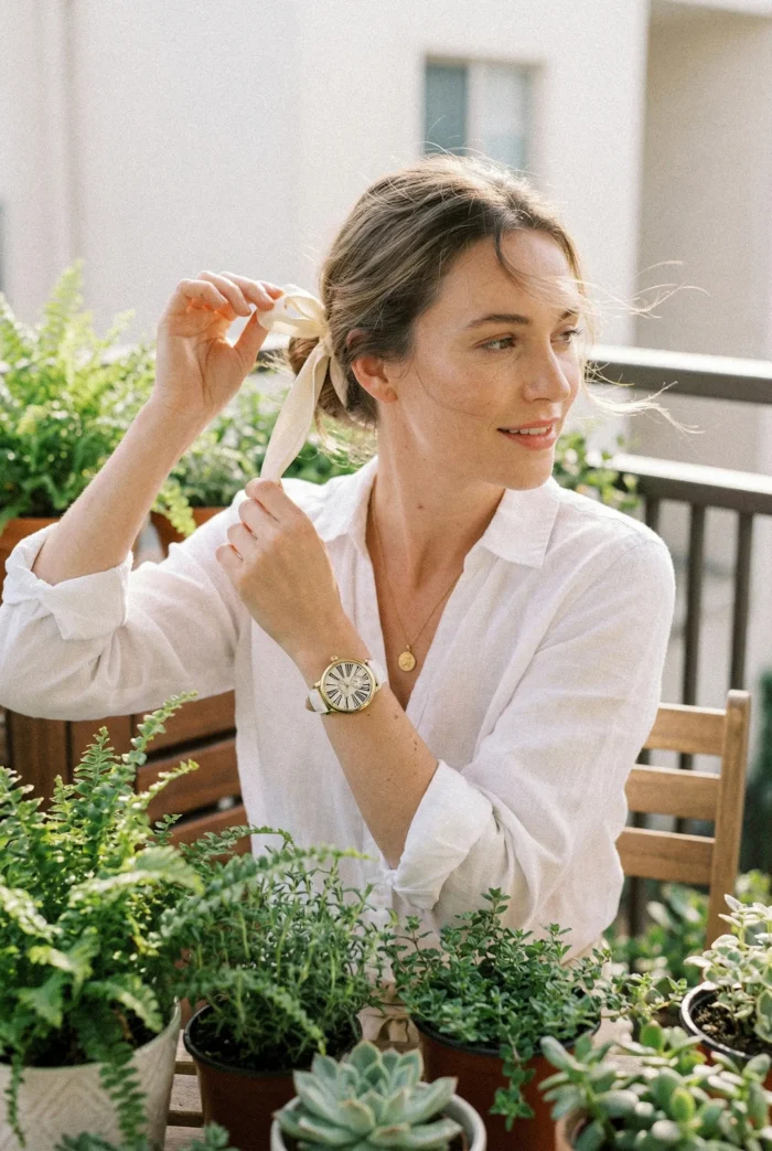 A woman in a white blouse ties her hair with a ribbon on a balcony. She's surrounded by green plants, exuding a calm and serene atmosphere.