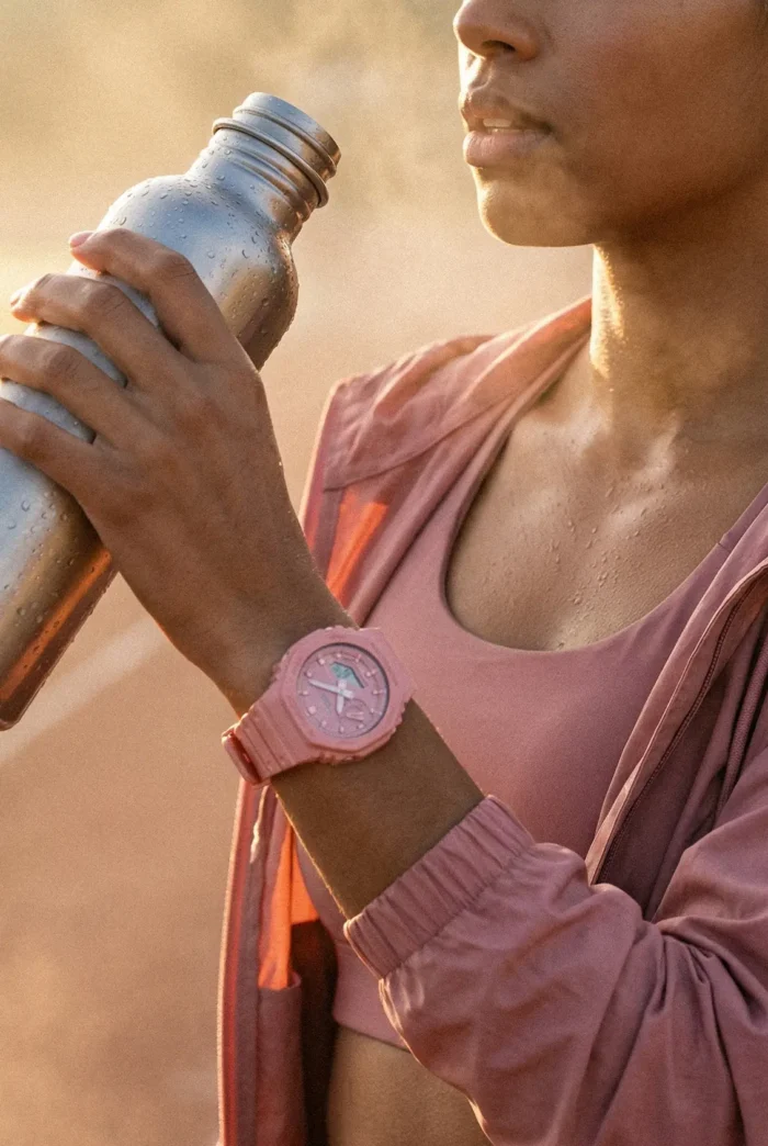 A woman in a pink sports outfit drinks from a metallic water bottle. She wears a pink watch and has a focused expression, with a warm, outdoor background.