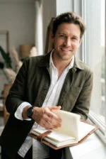 Man in an olive jacket and white shirt smiles while flipping through a notebook near a window. The space feels bright and welcoming.