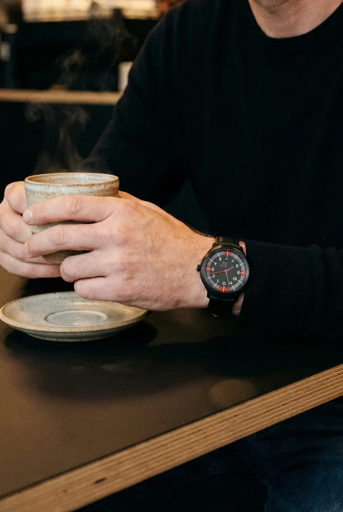 A person in a black sweater holds a steaming ceramic mug at a café table. They wear a black watch with a red and gray face, conveying a relaxed tone.