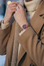 A person wearing a cozy beige coat and a knitted scarf adjusts their wristwatch with a burgundy face and gold accents. The setting feels warm and elegant.