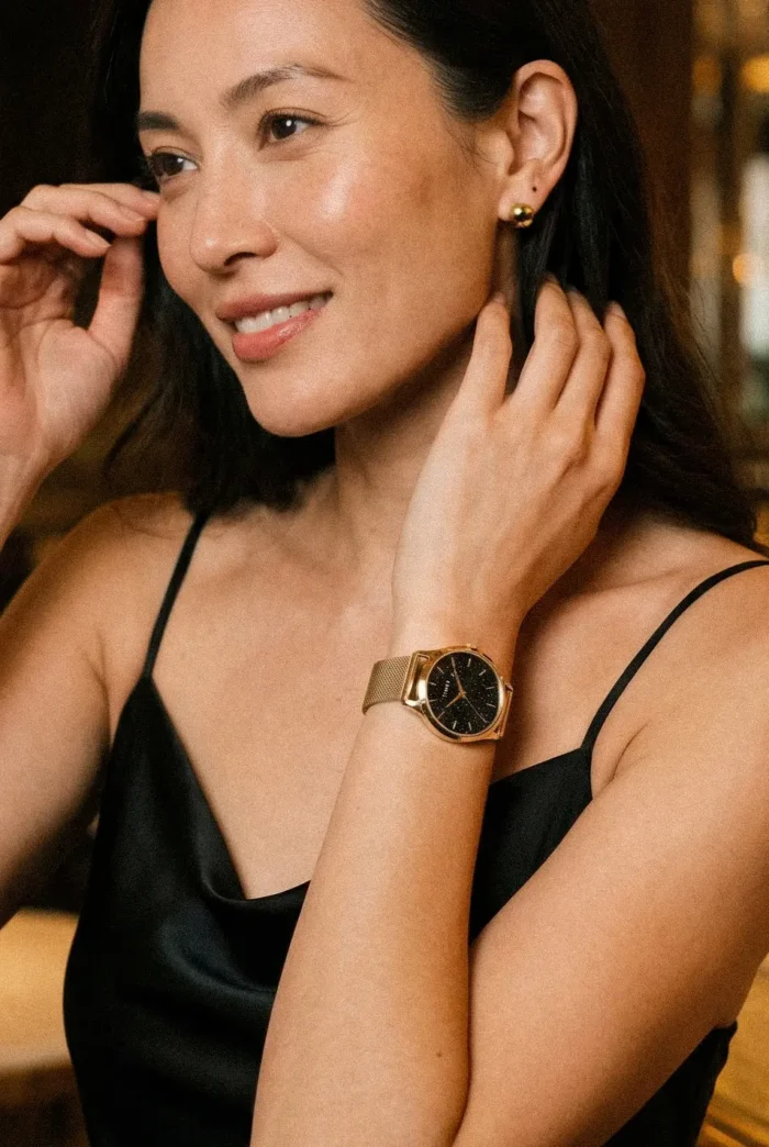 A close-up of a woman wearing a black satin dress, featuring a stylish gold watch and elegant earrings, with her hair down.