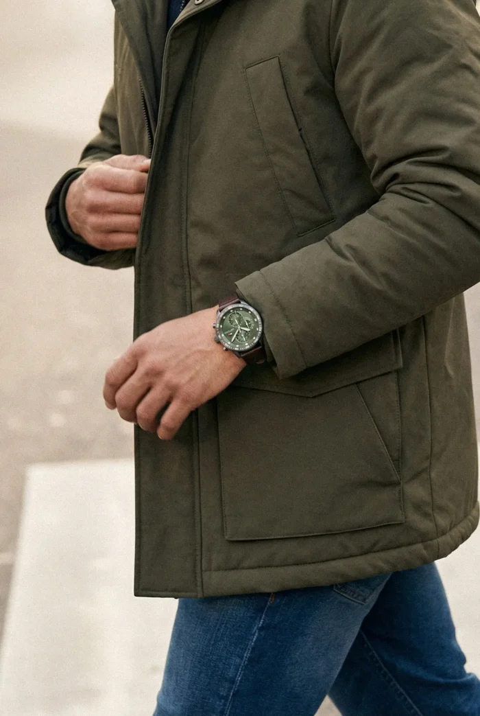 A person wearing a green coat and blue jeans walks, displaying a stylish green-faced wristwatch. The tone is casual and trendy.