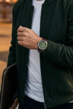 A person in a dark green jacket and white shirt is shown wearing a green-faced watch with a brown strap, holding a laptop case. The tone is stylish and casual.