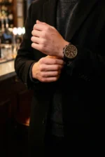 A person in a black coat adjusts their brown watch, standing in a dimly lit, elegant bar. The scene conveys sophistication and style.