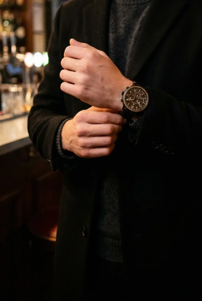 A person in a black coat adjusts their brown watch, standing in a dimly lit, elegant bar. The scene conveys sophistication and style.