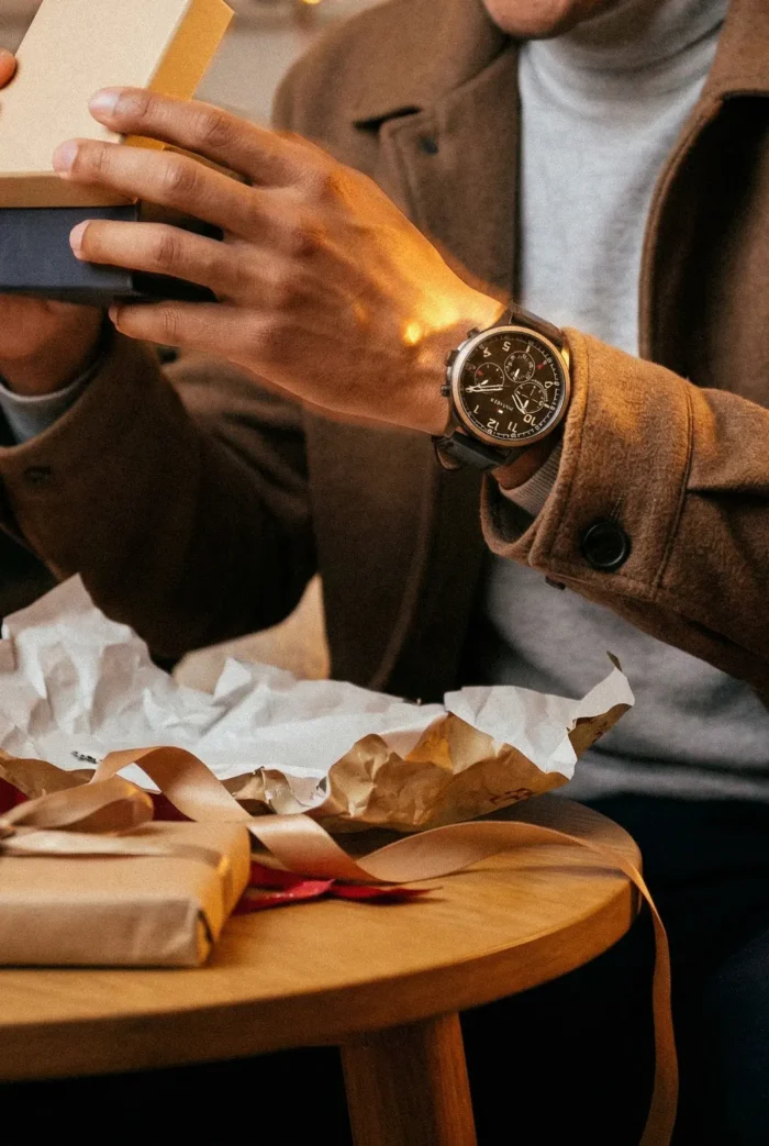 A person in a brown coat and watch is opening a gift box at a wooden table. Wrapping paper and ribbon are scattered, creating a warm, festive atmosphere.