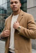 A man wearing a tan coat and cream sweater stands on an urban street, exuding confidence. He adjusts his golden watch, conveying a sense of sophistication.