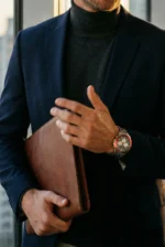 A person wearing a dark turtleneck and blazer holds a brown leather folder. They wear a watch with a red bezel, standing in front of a window.