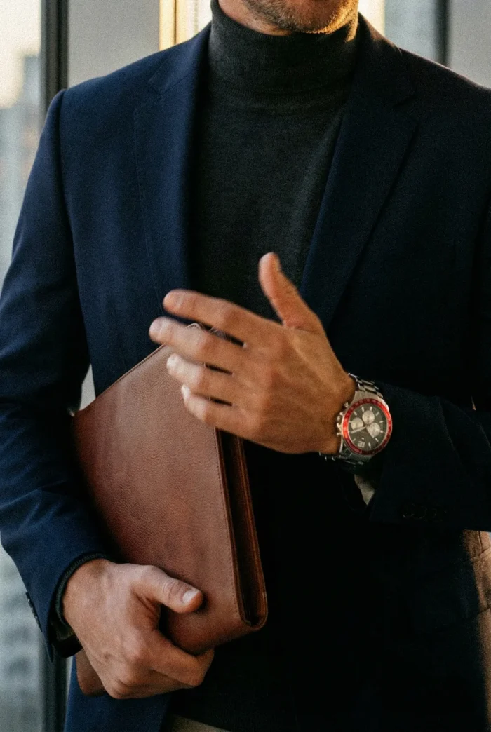 A person wearing a dark turtleneck and blazer holds a brown leather folder. They wear a watch with a red bezel, standing in front of a window.