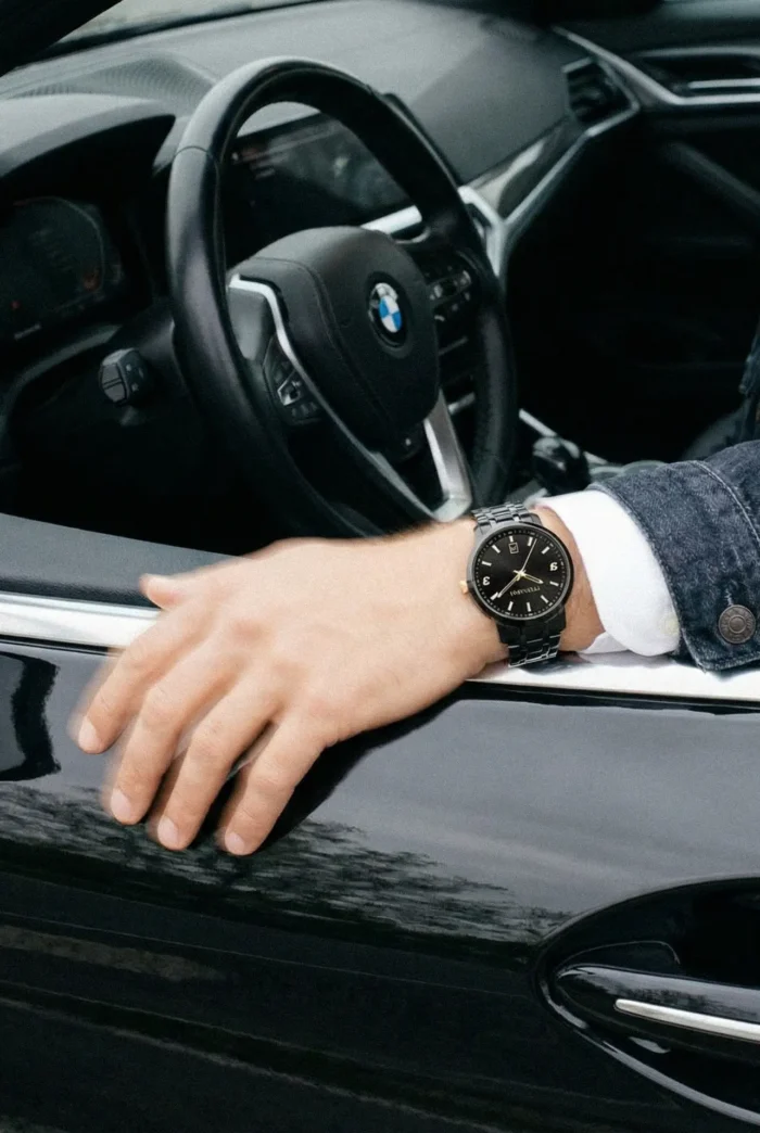 Man wearing a black watch rests his hand on the open window of a black BMW. The interior is partly visible, conveying a sense of luxury and style.