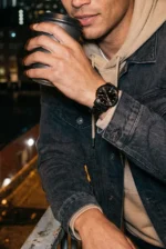 Man in a denim jacket and beige hoodie holding a coffee cup on a balcony at night, wearing a black watch, conveying a relaxed urban vibe.