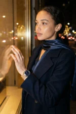 A woman in a dark coat and scarf gazes pensively through a brightly lit shop window at night. City lights create a warm and reflective ambiance.