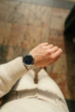 A person in a cream sweater and pants checks the time on a silver watch with a dark blue face. The mood is casual, standing on a tiled floor.