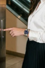 A woman in a white blouse and black skirt is pressing an elevator button. She wears a stylish blue-faced wristwatch, conveying a business casual tone.