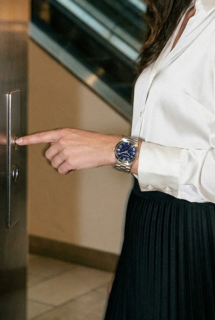 A woman in a white blouse and black skirt is pressing an elevator button. She wears a stylish blue-faced wristwatch, conveying a business casual tone.