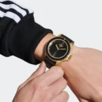 A close-up of a hand wearing a black and gold watch, with an Adidas three-stripe sleeve visible. The background is plain white.