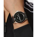 A close-up of a sleek black wristwatch with a round face, set against a person's arm, wearing a black zip-up jacket.