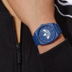 A blue sports watch with a minimalist design features the Adidas logo, worn on a wrist against a black zip-up jacket.