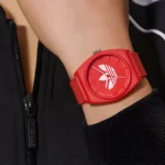 A close-up of a red wristwatch with a bold logo, worn on a person’s arm, paired with a black zip-up jacket.