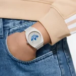 A close-up of a person's wrist wearing a stylish white watch with a blue design, alongside denim jeans and a light beige sweatshirt.