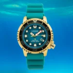 Auksinis ir turkio spalvos „Citizen Eco-Drive“ narų laikrodis su mėlynu ciferblatu, atsparus vandeniui iki 200 metrų, rodomas povandeniniame fone.