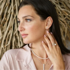 A woman with long dark hair wears delicate pearl earrings and layered necklaces, showcasing elegant jewelry against a textured backdrop.