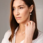 A woman wears elegant pearl jewelry, including a necklace, earrings, and a ring, against a neutral, softly lit background.