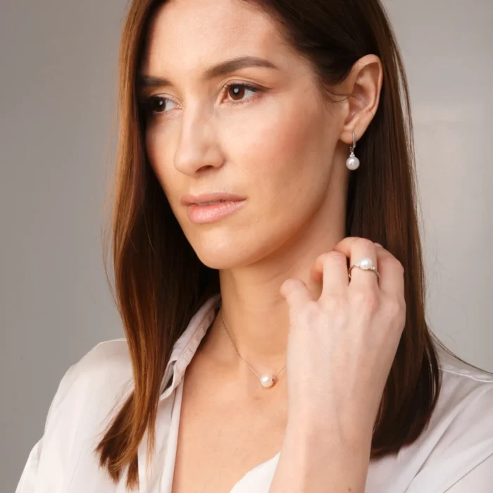 A woman wears elegant pearl jewelry, including a necklace, earrings, and a ring, against a neutral, softly lit background.