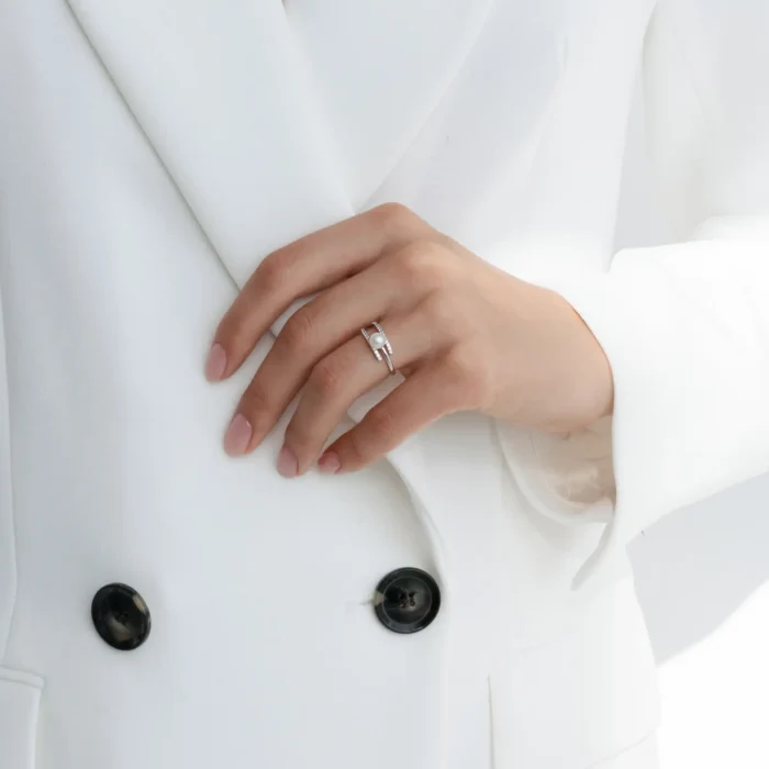A close-up of a hand wearing a silver ring with a pearl, resting on a white blazer with black buttons. Elegant and minimalist style