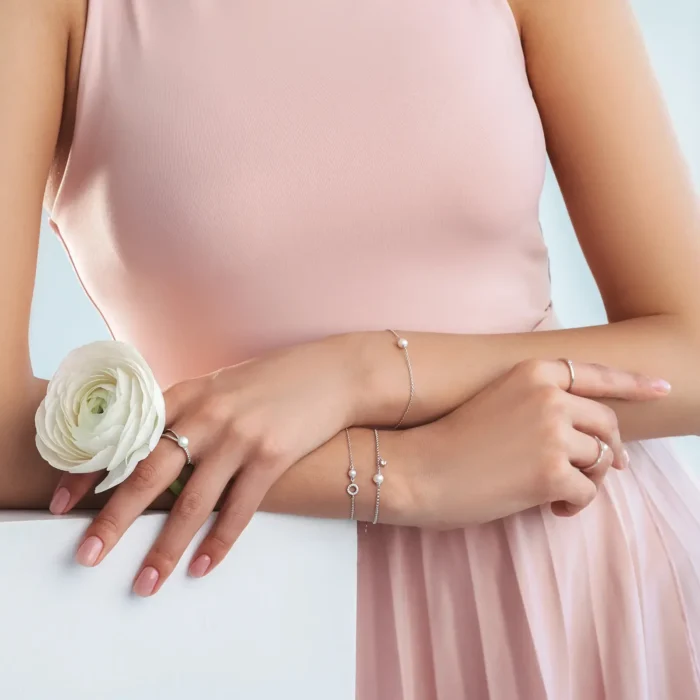 A woman in a pink dress showcases elegant jewelry, holding a white flower, with delicate rings and bracelets accentuating her graceful hands.