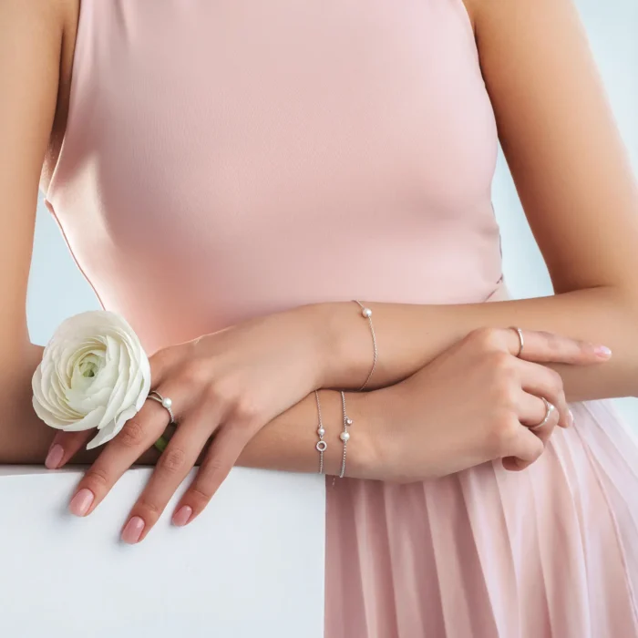 A woman in a pink dress showcases elegant jewelry, holding a white flower, with delicate rings and bracelets accentuating her graceful hands.