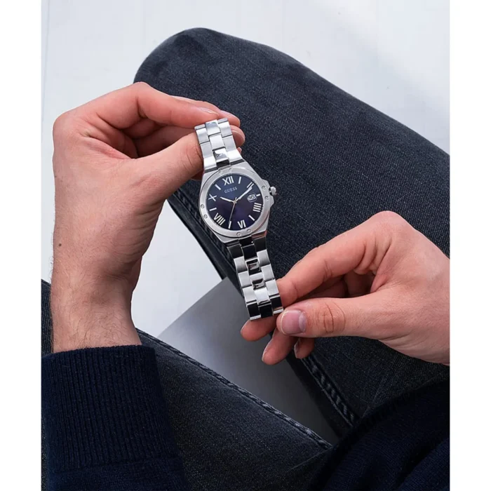 A person holding a silver Guess watch with a dark blue face, Roman numerals, and a stainless steel band against a stylish backdrop.